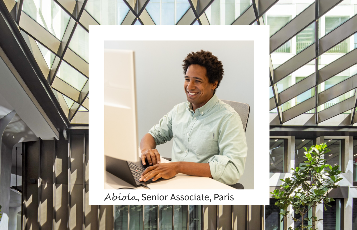 The image prominently features a white frame that encloses a photograph of Abiola, a Senior Associate in Paris. Within this frame, Abiola is seen sitting at a desk, smiling as he works on a computer. He wears a light green button-up shirt, conveying a professional yet approachable demeanor. The modern office interior in the background showcases geometric patterns on the windows and includes a touch of greenery with a potted plant visible at the bottom. Below the photo, his name and title, "Abiola, Senior Associate, Paris," are written in a clean, black font, emphasizing his role and location.
